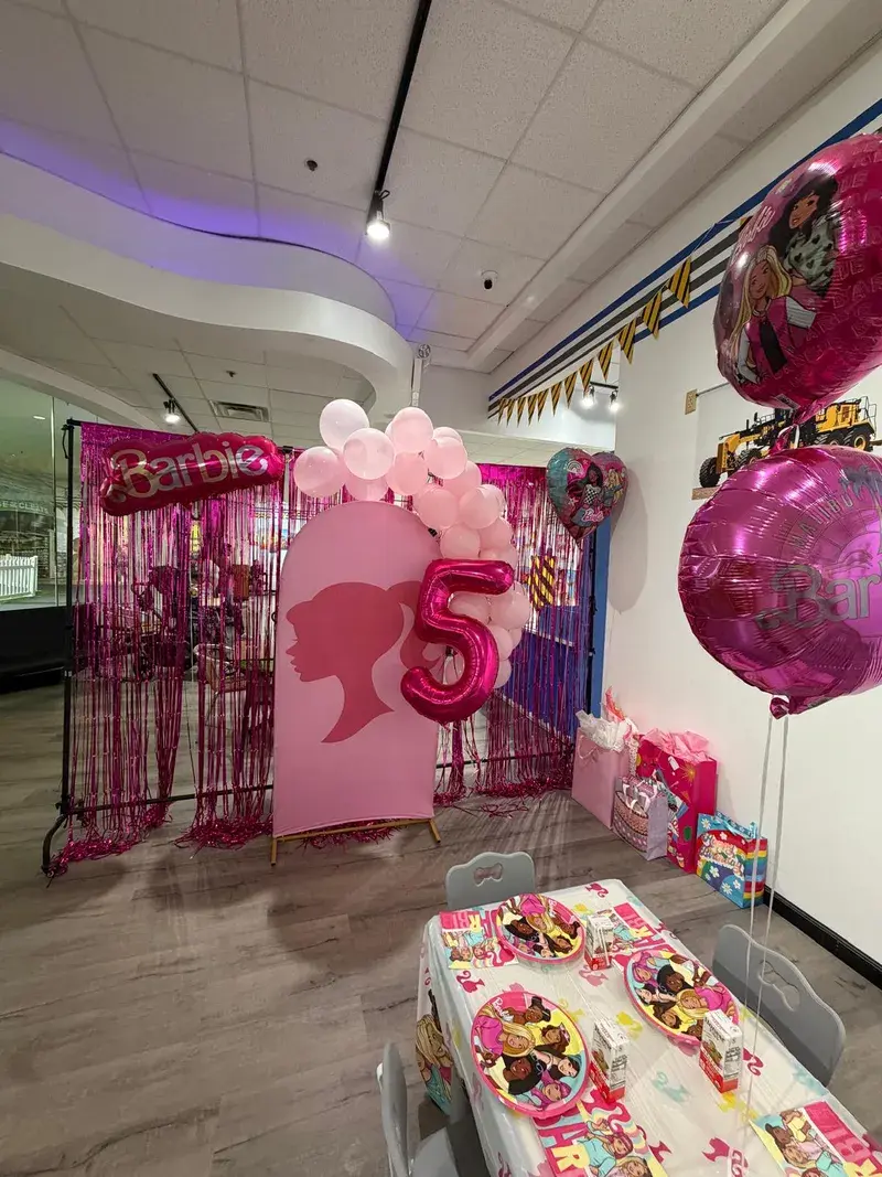 The party room decorated for a Barbie birthday, with a pink backdrop, balloon garland and a themed cake table