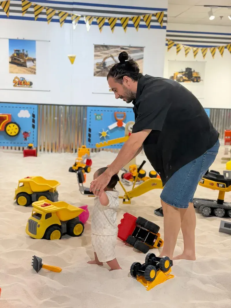A parent steadying a toddler beside toy dump trucks